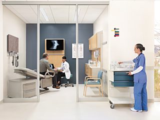 doctor interacting with patient in patient room. staff member in blue scrubs on right hand side, gathering supplies.