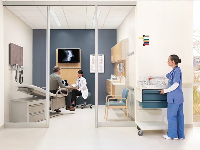 doctor interacting with patient in patient room. staff member in blue scrubs on right hand side, gathering supplies.
