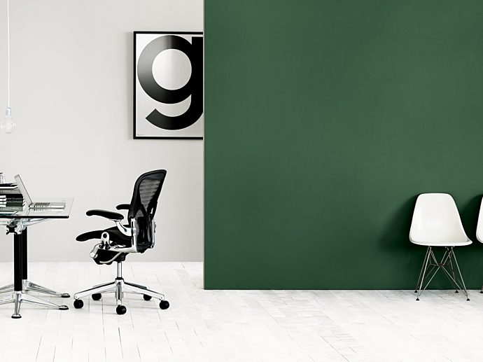 Left hand side of room with white walls with a glass desk and black office chair. The right is two white Eames Molded Plastic chair against green wall.