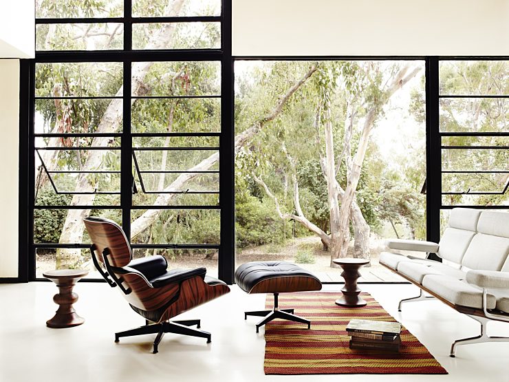 Classic Eames Lounge and ottoman paired in a modern livingroom with walnut stools an Eames sofa with a large picture window in the background.