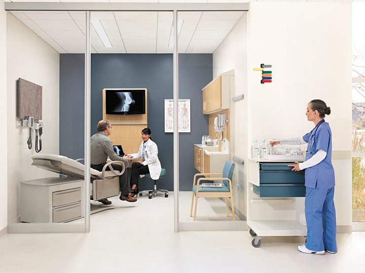 doctor interacting with patient in patient room. staff member in blue scrubs on right hand side, gathering supplies.